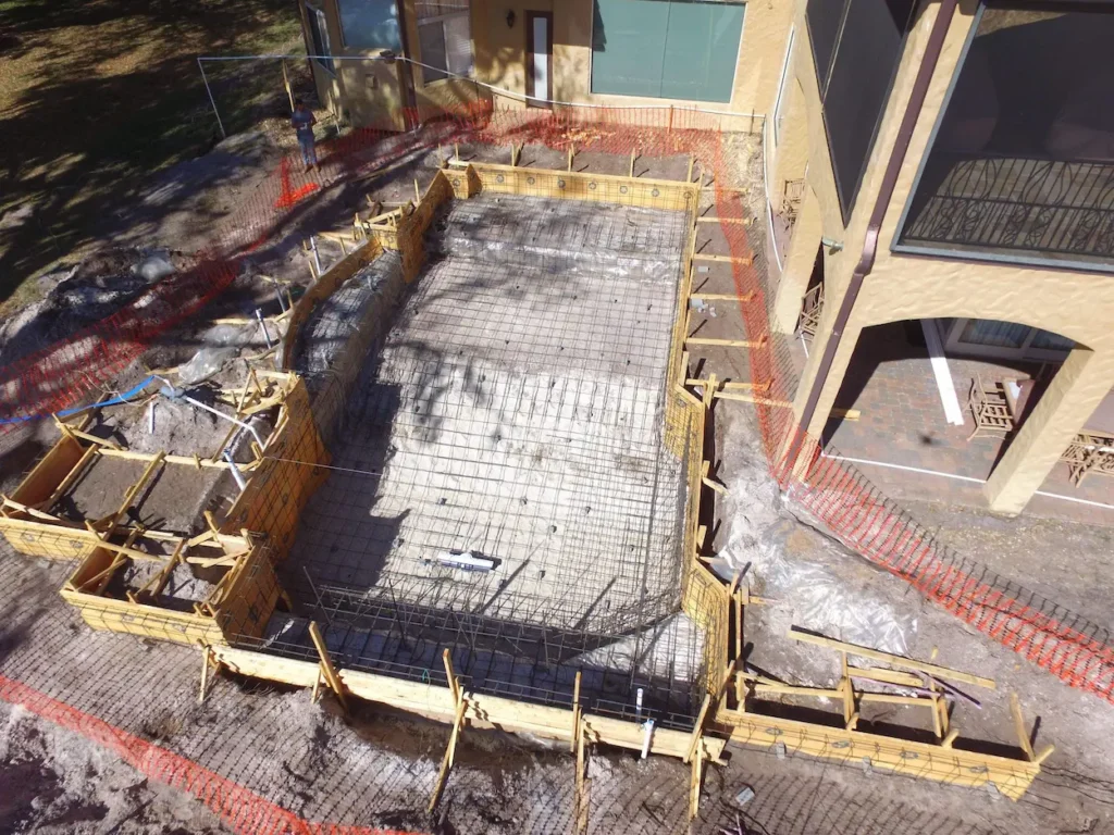 A rectangular in-ground pool is under construction in a backyard, with rebar grid laid and wooden framework surrounding the area next to a tan house. Orange safety fencing is set up around the site, highlighting the early stages of gunite only contracting.